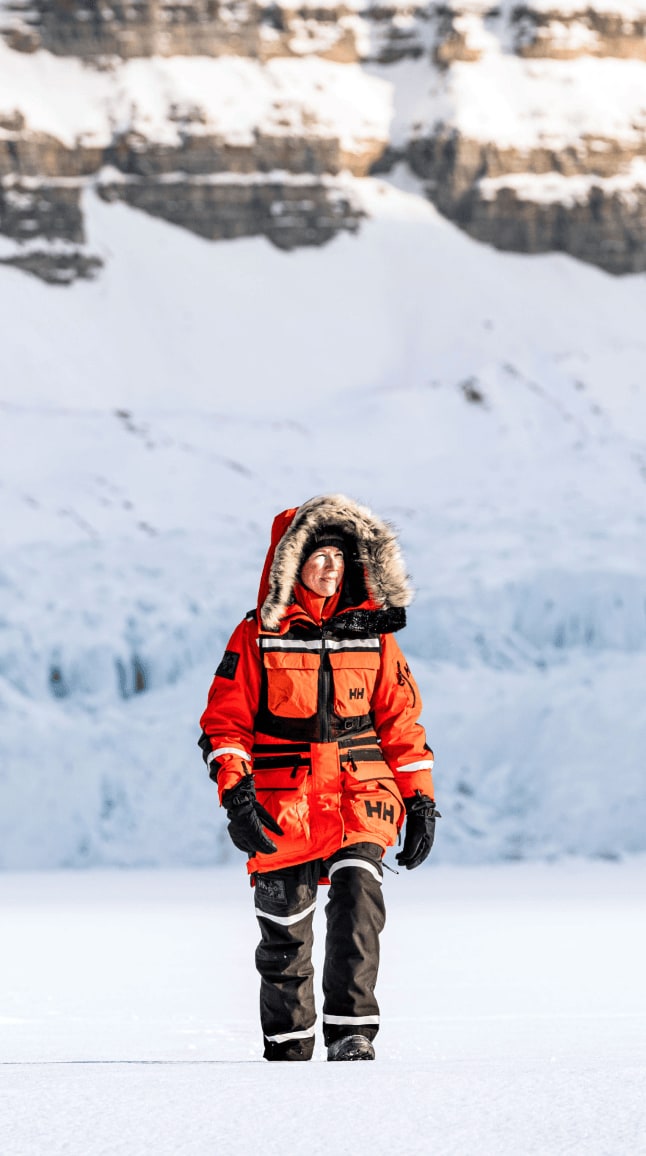 Heïdi marche sur une surface gelée, entourée d'un paysage glaciaire monumental. Elle porte une épaisse parka orange bordée de fourrure de la marque HH, des gants noirs et un pantalon technique. Derrière elle, on distingue la paroi bleutée d'un glacier. Son regard est tourné vers l'horizon, reflétant la détermination et la sérénité d'une exploratrice évoluant dans les conditions extrêmes de l'Arctique.