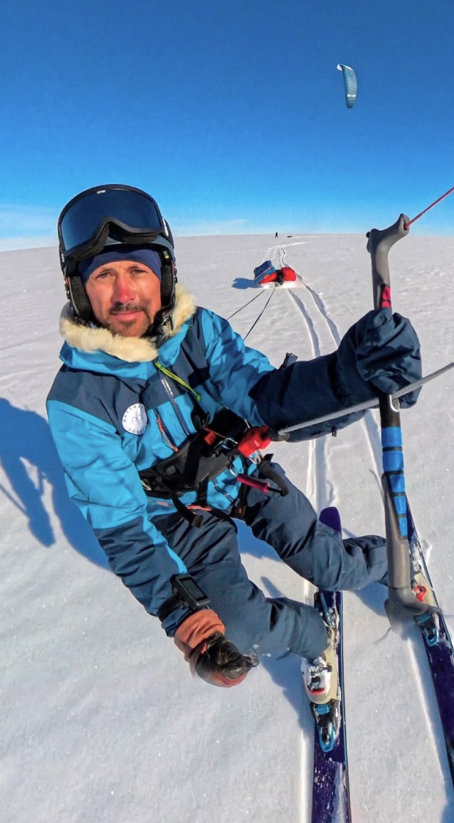 Matthieu pratique le snowkite sur une vaste plaine enneigée. Penché légèrement vers la gauche, les genoux fléchis sur ses skis, il tient fermement la barre de son aile de traction. Sa pulka rouge est tractée derrière lui sur la neige. Il porte une combinaison bleue, un bonnet bleu et un casque sur lequel repose son masque de ski. Son visage exprime un léger sourire. Le ciel est d'un bleu éclatant, sans nuage, et la lumière intense accentue la blancheur du paysage. Au loin, on distingue Heïdi, plus petite dans la perspective, avançant elle aussi sur la neige. L'image transmet une sensation de mouvement, de vitesse et de liberté au cœur d'un environnement polaire infini.