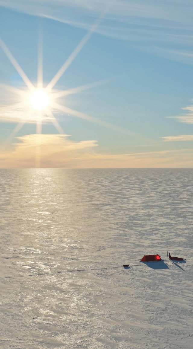 Sous un soleil éclatant rasant l'horizon, une tente rouge se dresse au milieu de l'immensité blanche de la calotte polaire. À ses côtés, du matériel d'expédition est aligné sur la neige, projetant de longues ombres sur le sol glacé. L'horizon se perd dans une lumière dorée où le ciel bleu pâle se fond dans les reflets irisés des nuages. Cette scène saisit la solitude et la beauté silencieuse d'un bivouac au cœur de l'Antarctique.