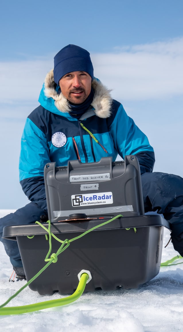 Matthieu est assis sur la neige, au cœur d'un vaste paysage glacé qui s'étend à perte de vue sous un ciel bleu clair légèrement voilé de nuages. Il porte une veste d'expédition bleue et turquoise à capuche bordée de fourrure, un bonnet bleu marine et un pantalon technique isolant. Devant lui, une grande caisse noire portant l'étiquette IceRadar – bluesystem.ca contient le matériel scientifique utilisé pour leurs relevés de la calotte polaire. Une corde verte relie la caisse à d'autres instruments étendus sur la glace, visibles en arrière-plan. Matthieu regarde vers la droite de l'image, le visage concentré et légèrement éclairé par la lumière du soleil arctique. L'ensemble évoque la rigueur et la précision du travail scientifique mené dans des conditions extrêmes.