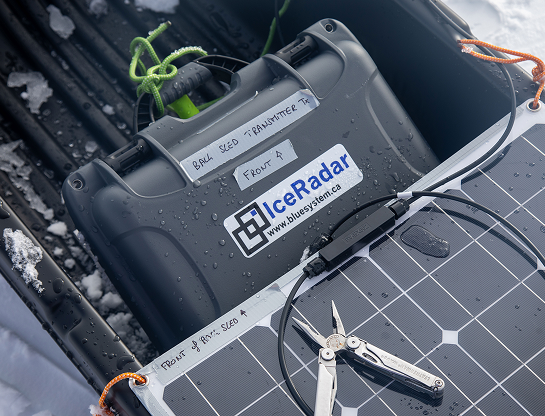 Gros plan sur le matériel scientifique de l'expédition. À l'intérieur d'une pulka noire, on distingue un boîtier gris foncé étiqueté IceRadar – Blue System, utilisé pour les mesures de profondeur de la glace. Des étiquettes manuscrites précisent sa position sur le traîneau et sa fonction de « transmitter ». Un panneau solaire repose sur la pulka, relié par un câble d'alimentation, tandis qu'un outil multifonction en acier est posé à proximité. Des gouttes de glace et de condensation témoignent du froid ambiant.