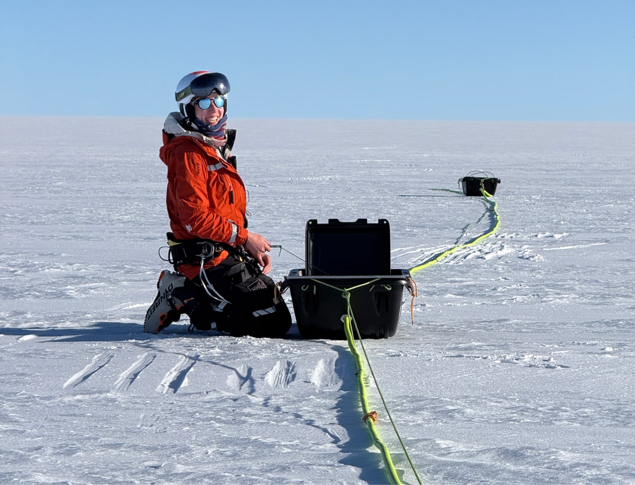 Sur la calotte glaciaire, Heïdi est agenouillée dans la neige, souriante face à la caméra. Elle porte une veste orange vif, un casque et des lunettes de ski, avec un baudrier d'expédition autour de la taille. Devant elle, une mallette scientifique est ouverte, reliée à un long câble jaune fluorescent qui serpente sur la glace jusqu'à une autre caisse plus loin. La lumière met en valeur la blancheur du paysage et l'énergie positive du moment.