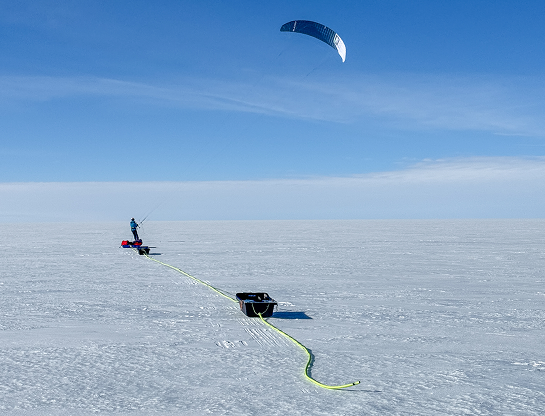 Sur la calotte glaciaire, Matthieu avance en snowkite, tracté par une grande aile bleue et blanche qui se détache dans le ciel. Il tire derrière lui une pulka rouge et deux radars scientifiques noirs reliés par un câble jaune fluorescent serpentant sur la glace. La scène illustre à la fois la dimension sportive et scientifique de l'expédition : Matthieu se déplace à la force du vent tout en réalisant des relevés géophysiques. L'horizon est parfaitement dégagé, baigné d'une lumière polaire éclatante.